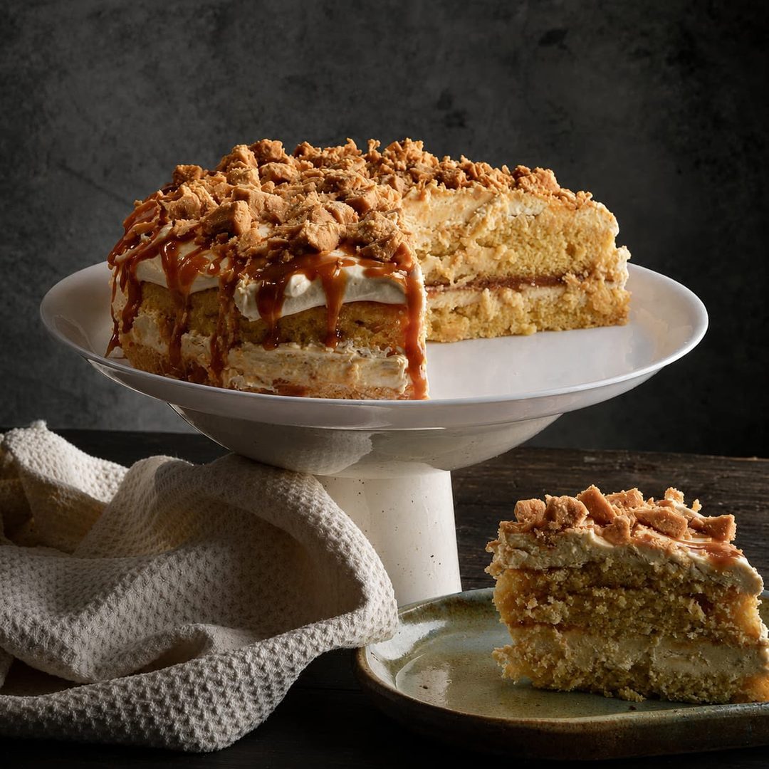 A caramel biscuit layer cake on a white stand, with a slice served on a ceramic plate